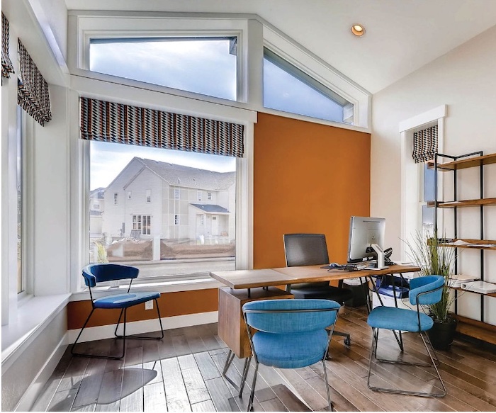 The office has an orange wall, large 100 Series windows showing a view of houses, a wooden desk, and two blue chairs.