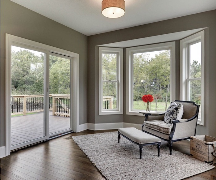 A cozy reading nook with a beige armchair and ottoman sits in front of a 200 series bay window overlooking a deck and wooded area. The room features hardwood floors, a large sliding glass door leading to the deck, and a neutral color palette. A fluffy, light-grey rug sits under the seating arrangement.