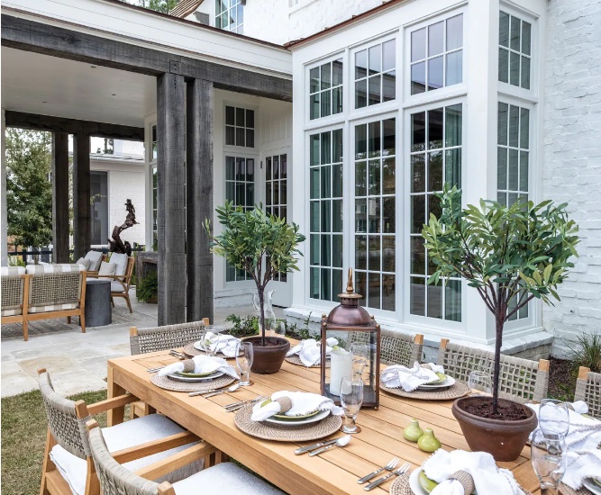 Outdoor dining scene: a long wooden table with place settings is surrounded by patio chairs.  Potted plants and a white house with A Series Windows are visible in the background.