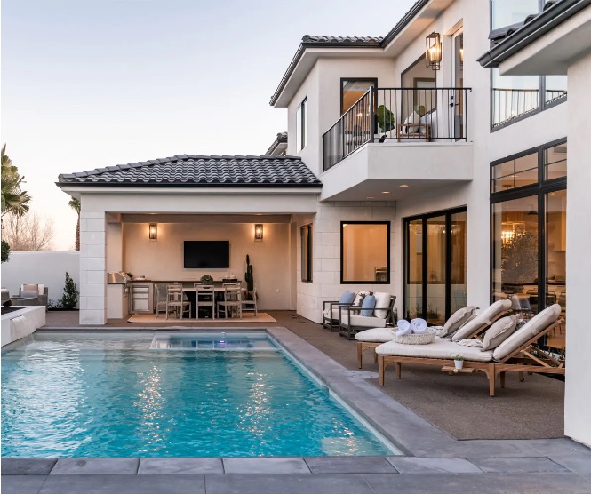 A luxurious backyard features a rectangular swimming pool, an outdoor kitchen under a covered patio, and comfortable lounge chairs surrounding the pool. The house is modern in style, with white stucco walls and large windows and doors.