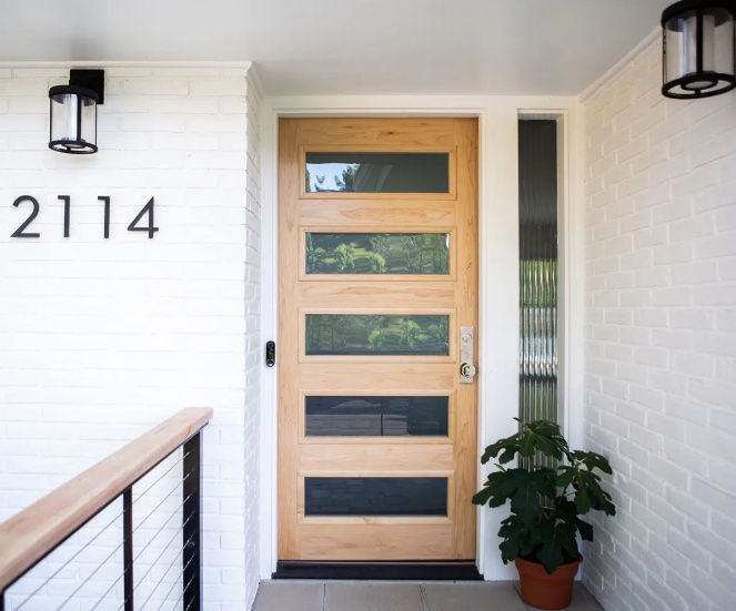 A modern home's front entrance features a light wood door with horizontal glass panels. The door is set within a white brick wall, accented by a sleek, black house number "2114" and simple light fixtures. A small plant sits beside the door.