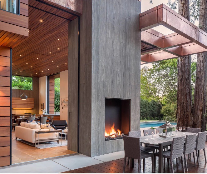 A sleek, modern home features an outdoor patio with a large stone fireplace, a dining table and chairs for al fresco dining, and a view of a swimming pool beyond.  The architecture includes wood and metal accents and hidden doors to bring the outdoors inside.