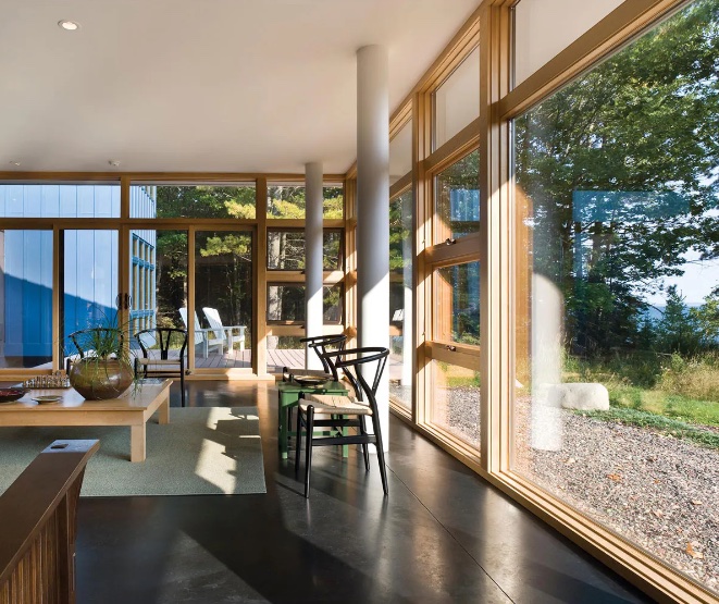 Interior shot of a bright, airy room with e series picture windows offering a view of nature.  The floor is dark and polished.  There are several chairs and a table.