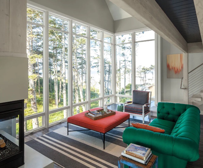 "A bright, airy living room with floor-to-ceiling e series windows showcasing a lush view. A vibrant green sofa sits in the foreground, with a coral-colored ottoman in front of it. A gray armchair and a small side table are also visible.  A modern fireplace is partially visible on the left, and a striped rug anchors the seating area.