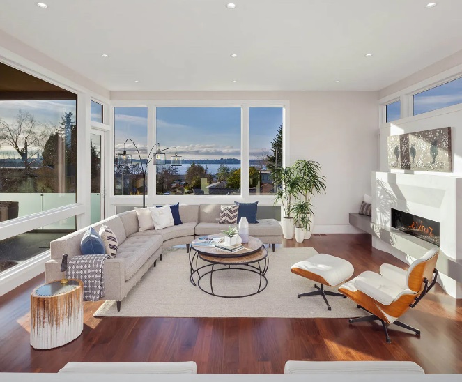 Spacious living room with a curved sofa, comfortable armchair, and a fireplace.  Large e series windows provide a beautiful view of a body of water and the horizon.